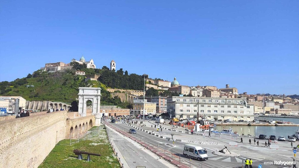 A coastal cityscape with a prominent hilltop fortress, a large open square, and a waterfront with boats. 1