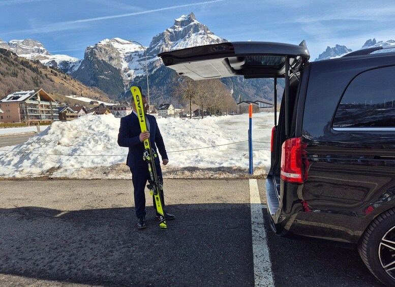 A person holding skis next to a black SUV with a snow-covered mountain in the background. 3