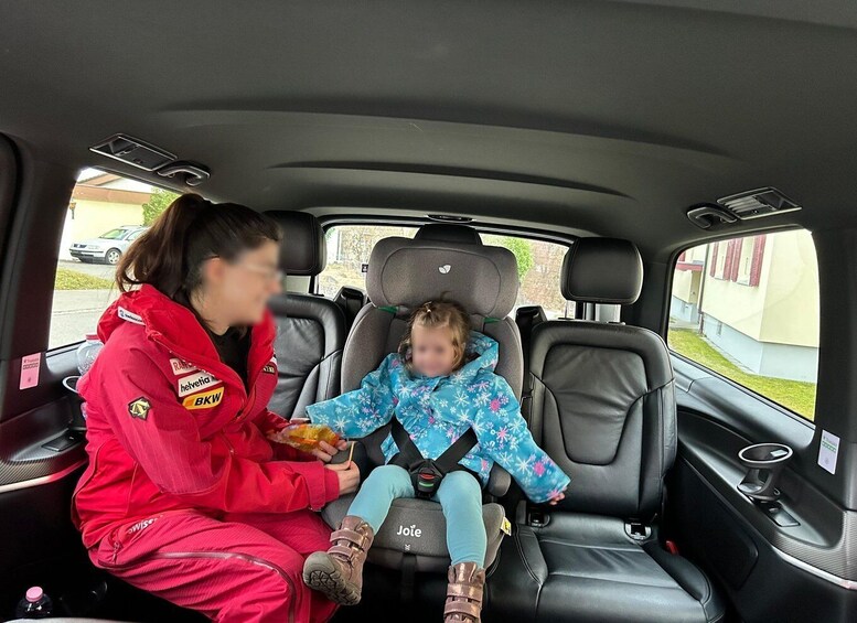 A woman in a red racing suit and a child in a blue snowflake-patterned coat seated in a car's back seat. 2