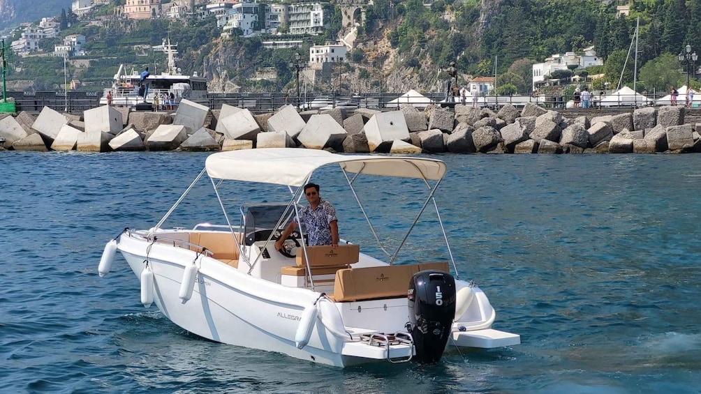 A small white motorboat with a canopy, a man steering, and a rocky shoreline with buildings in the background. 1