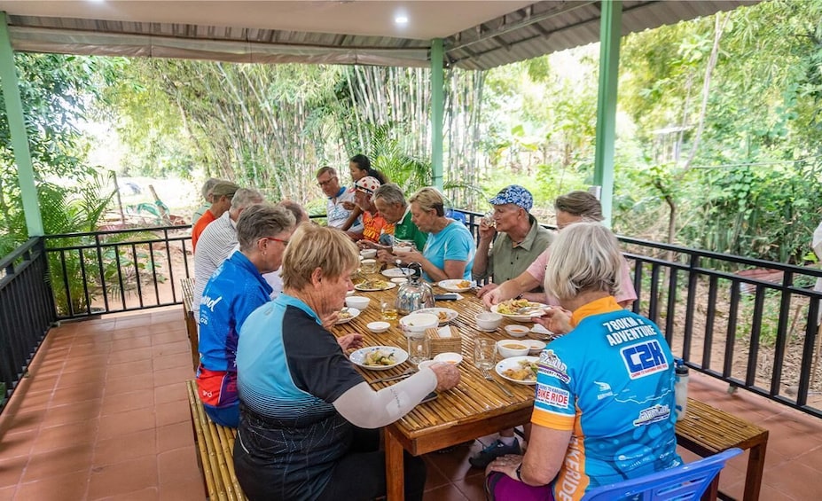A group of people dining outdoors at a covered table with bamboo mats, surrounded by lush greenery. 5