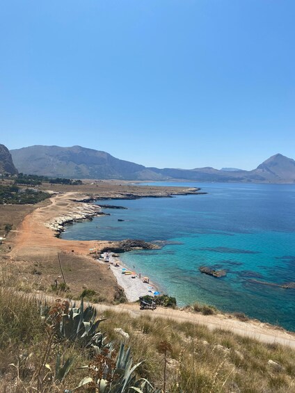 A coastal landscape with a dirt path, clear blue sea, rocky shoreline, and distant mountains. 1