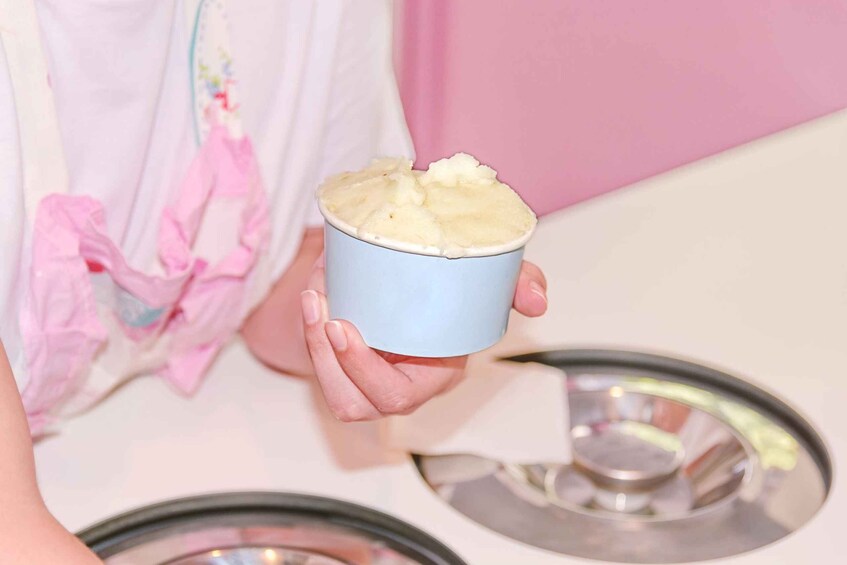 A person holding a blue cup with a white substance, possibly ice cream, in front of a pink wall. 1