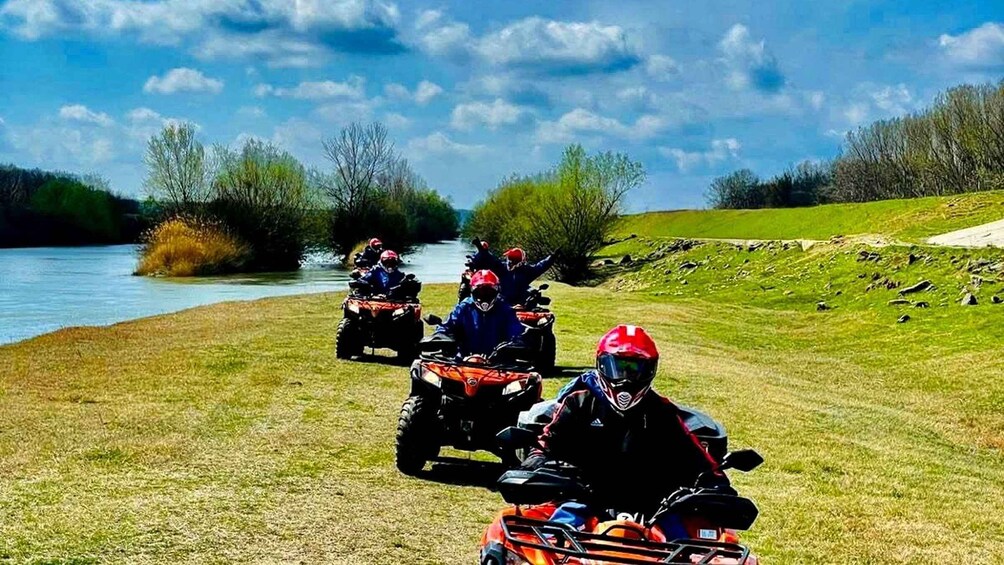 Four people on quad bikes near a river, with trees and a clear sky in the background. 1