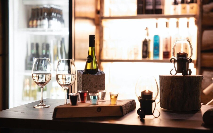 A bar setting with glasses of wine, a bottle of wine, shot glasses, and a candle on a wooden table. 3