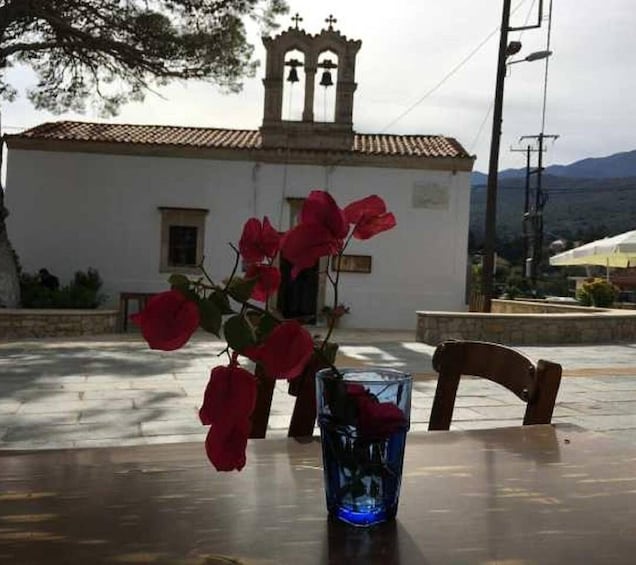 A table with a vase of red flowers and a glass of water, a chair, and a white building with a bell tower in the background. 5