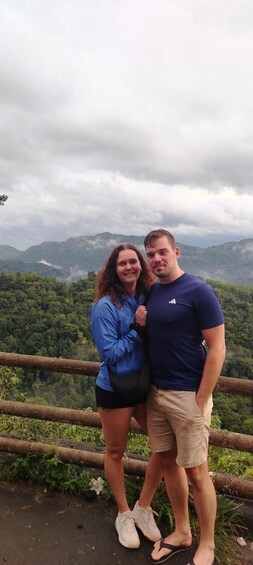 Dos personas de pie junto a una barandilla, con un paisaje montañoso al fondo. 3