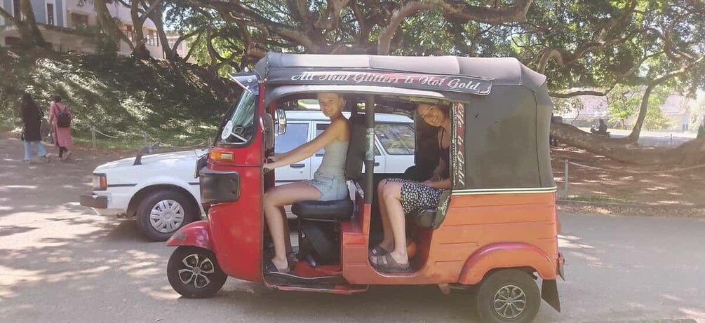 Un tuk-tuk rojo y naranja con pasajeros, estacionado en una calle con árboles y un coche blanco al fondo. 2