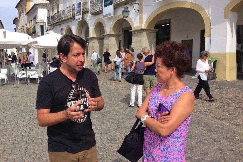Two people conversing in a cobblestone square with historic buildings in the background. 4