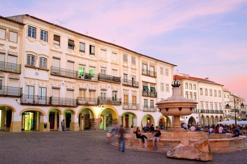 A historic square with a fountain, buildings with balconies, and people walking. 2