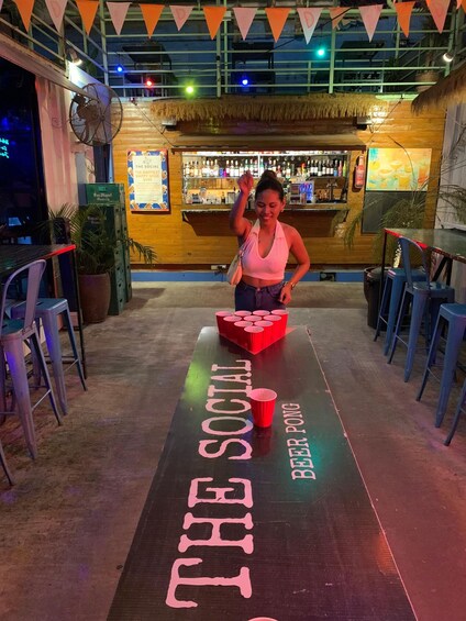 A bar with a table labeled 'The Social Beer Pong' and a woman standing behind it. 2