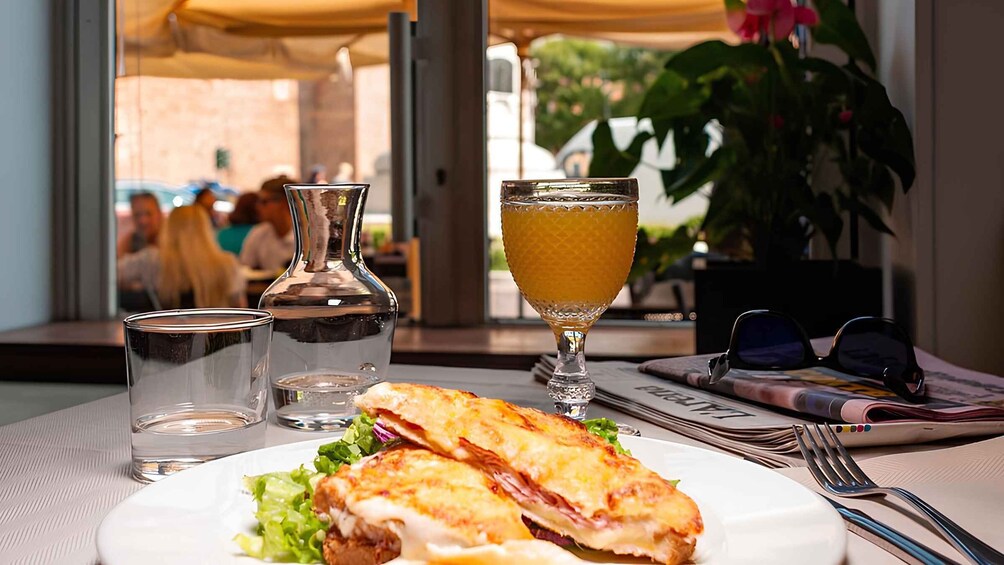 A sandwich on a plate, a glass of orange juice, a water glass, and a bottle of water on a table with a window view. 2