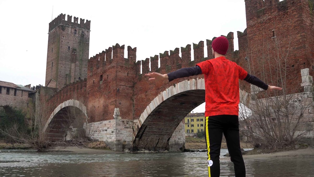 A person standing with arms outstretched in front of a medieval bridge with towers. 4