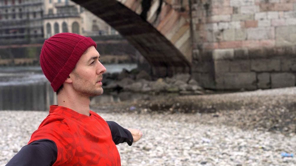 A person wearing a red beanie and a red vest, standing on a pebbled surface with a stone wall and a bridge in the background. 1