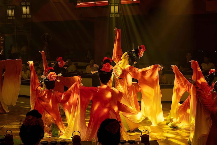 A group of dancers in traditional costumes performing on a stage with an audience seated in the background. 1