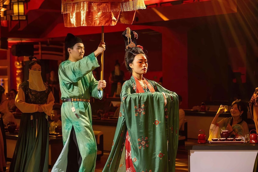 A person in traditional attire holding a fan, another in green with red patterns, and a table with red teapots and cups. 3