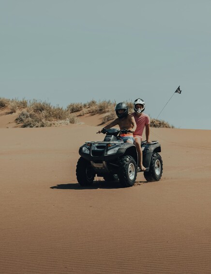 Two people on an ATV in a desert with sand dunes and sparse vegetation. 1
