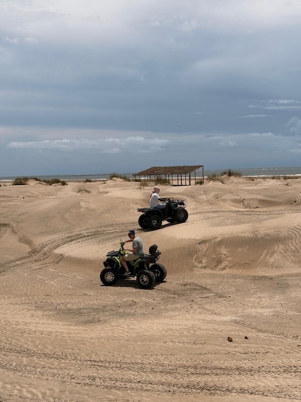 Two people on ATVs in a desert landscape with dunes and a shelter in the background. 3