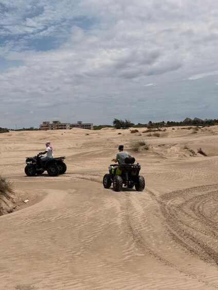 Two people riding ATVs on a sandy terrain with buildings in the background. 4