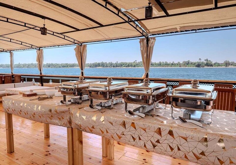 A buffet table with food service equipment on a boat deck with a view of a calm body of water and a distant shoreline. 3