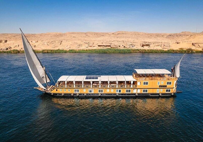 A yellow riverboat with sails on a calm river, surrounded by desert hills. 1
