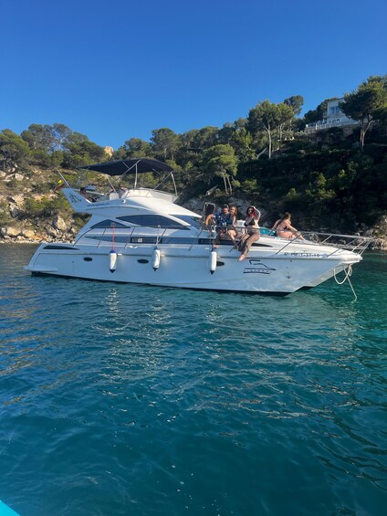 A white yacht with people on board, anchored near a rocky coastline with trees and a building in the background. 1
