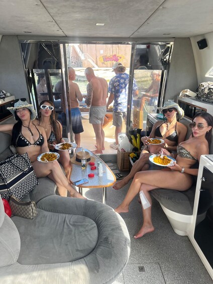A group of people on a boat, some eating, others standing, with a view of a dock and water. 1