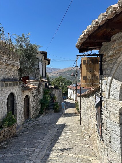 A narrow cobblestone street in a historic town with stone buildings, a sign for 'BEER & WINE,' and a person walking away. 2