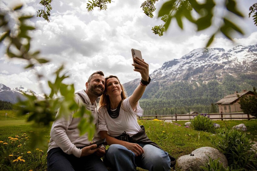 Two people taking a selfie in a mountainous landscape with a house in the background. 1
