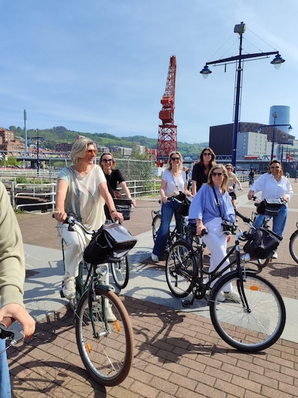 A group of people riding bicycles on a paved path with a red crane and industrial buildings in the background. 4