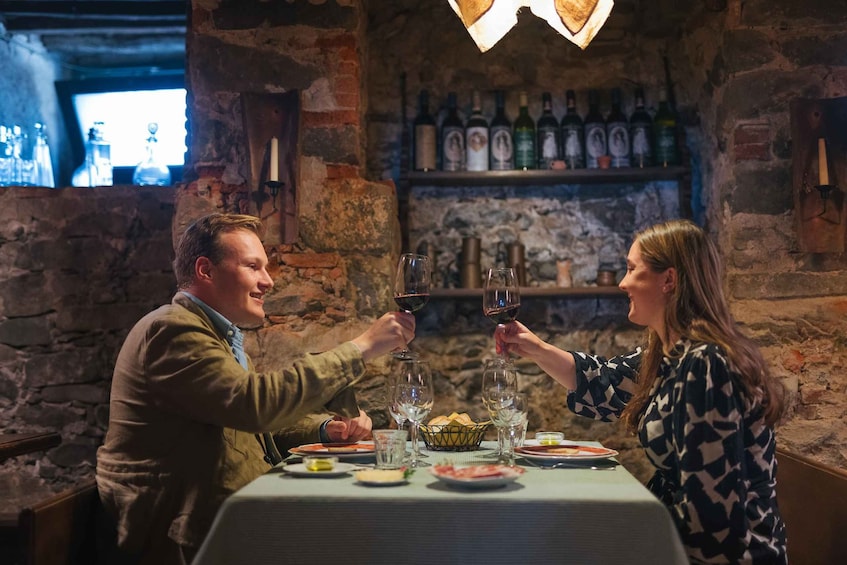 A couple toasting with wine glasses at a restaurant with a stone wall and a wine bottle shelf. 1