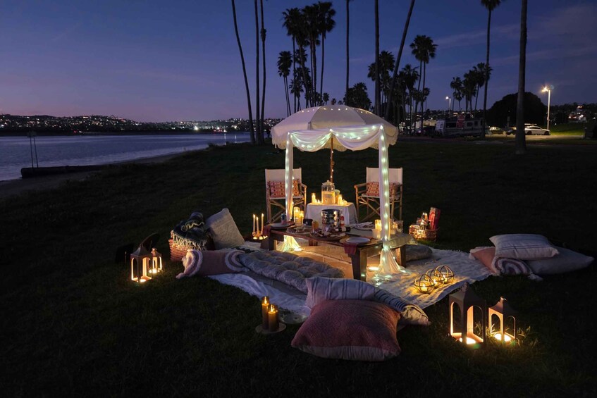 A picnic setup with a canopy, table, chairs, and lanterns on a grassy area by the water at dusk. 1
