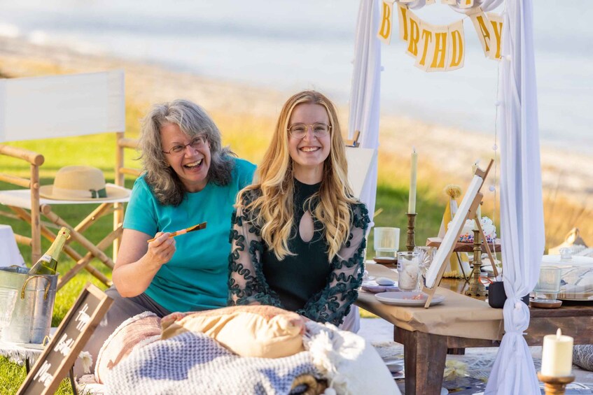 Two people sitting at a picnic table with a birthday banner, surrounded by art supplies and a scenic outdoor setting. 3
