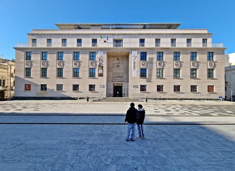 A large, modern building with a central entrance, multiple windows, and banners. 1