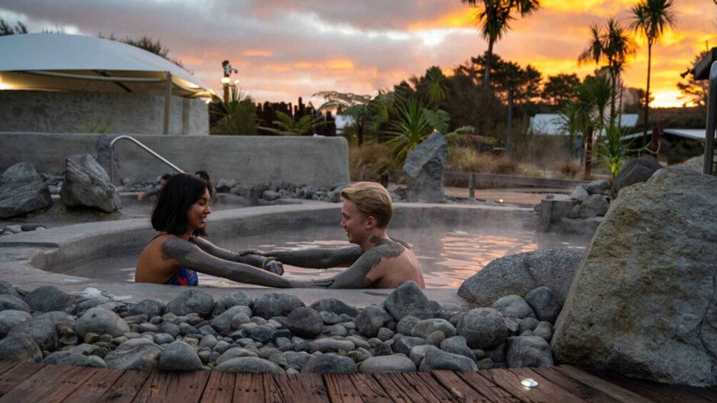 A couple in a hot spring surrounded by rocks and palm trees at sunset. 1
