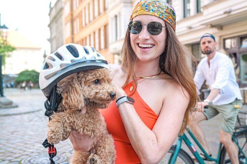 A person holding a poodle with a helmet, a bicycle, and buildings in the background. 5