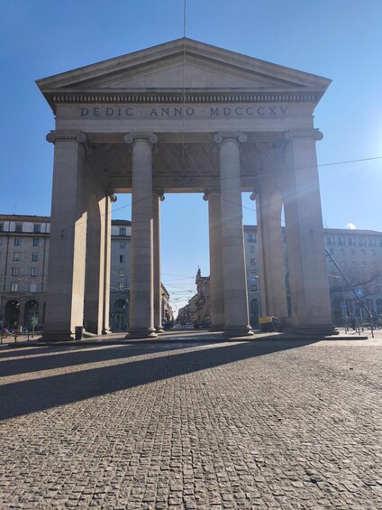 A neoclassical monument with inscriptions, surrounded by cobblestone pavement and urban buildings. 3