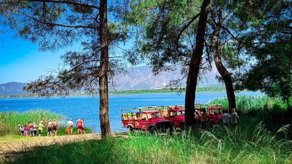 A group of people by a lake with a red vehicle and trees. 1