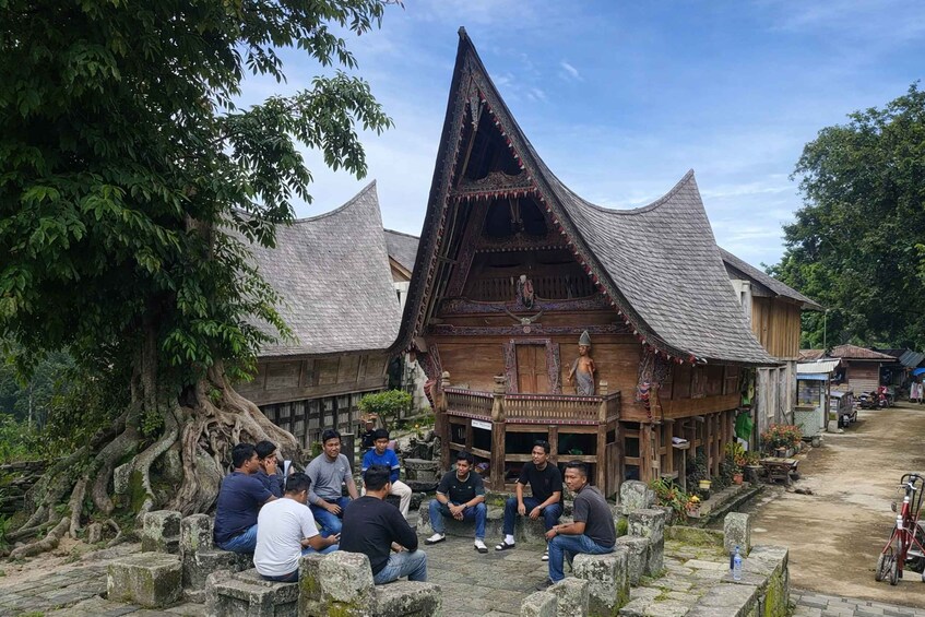 A group of people sitting on stone steps in front of a traditional wooden house with a thatched roof. 2
