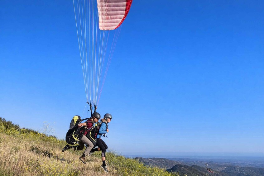 A person paragliding with a red and white parachute, running on a grassy hill with a clear blue sky. 1