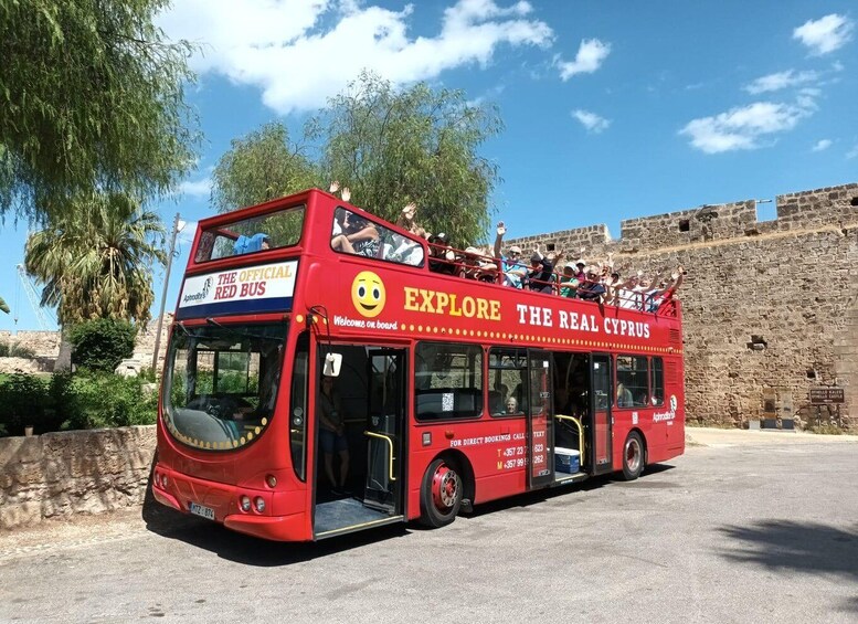 A red double-decker bus with 'EXPLORE THE REAL CYPRUS' written on the side, parked near a stone wall. 1