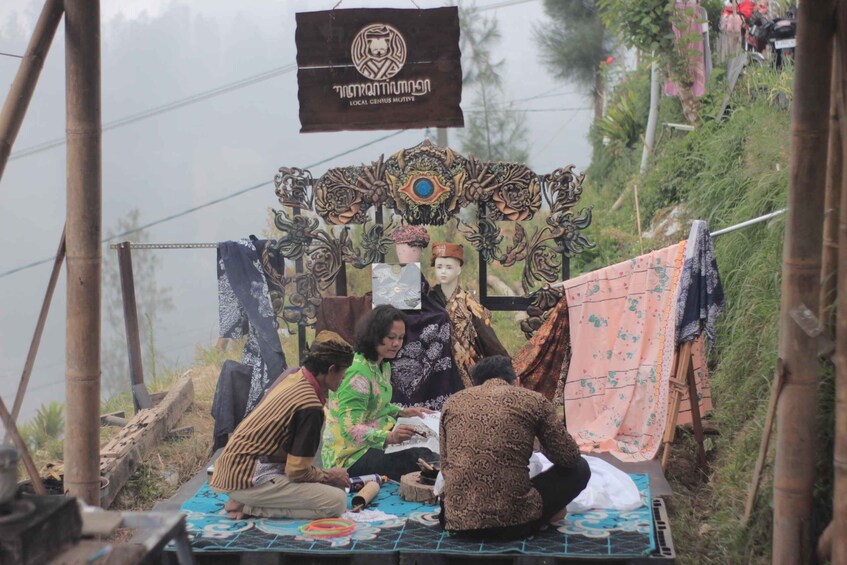A group of people sitting on a mat outdoors, surrounded by a decorative backdrop with a sign above. 2