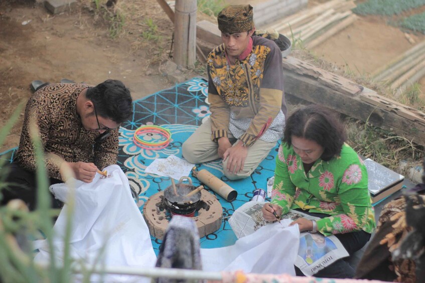 Three people sitting on a patterned mat outdoors, with one person writing on a piece of paper. 5