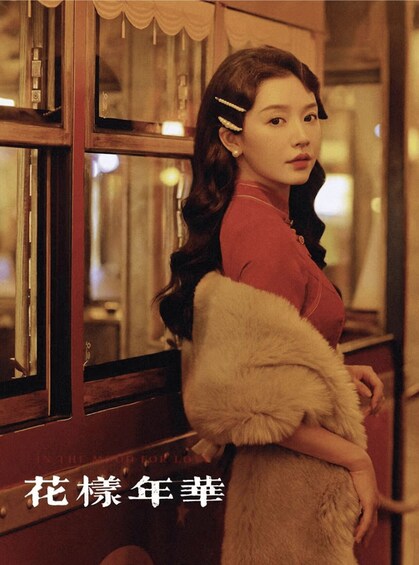A person in a red top and fur coat standing by a window with curtains. 5