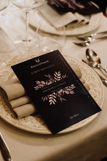 A black menu with 'Rocca d'Italia' and 'Summer Season Event' text, placed on a beige plate with a napkin. 5