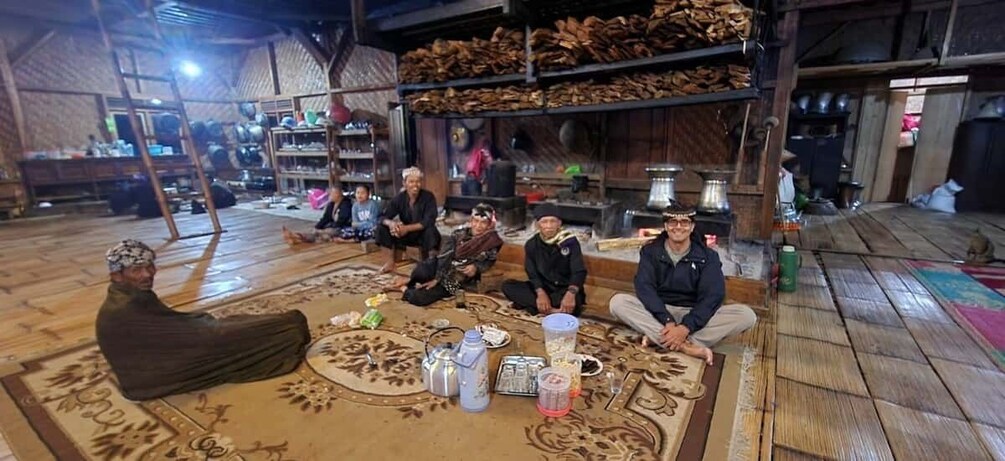 A group of people sitting on a patterned rug in a rustic wooden room with shelves of dried goods in the background. 4
