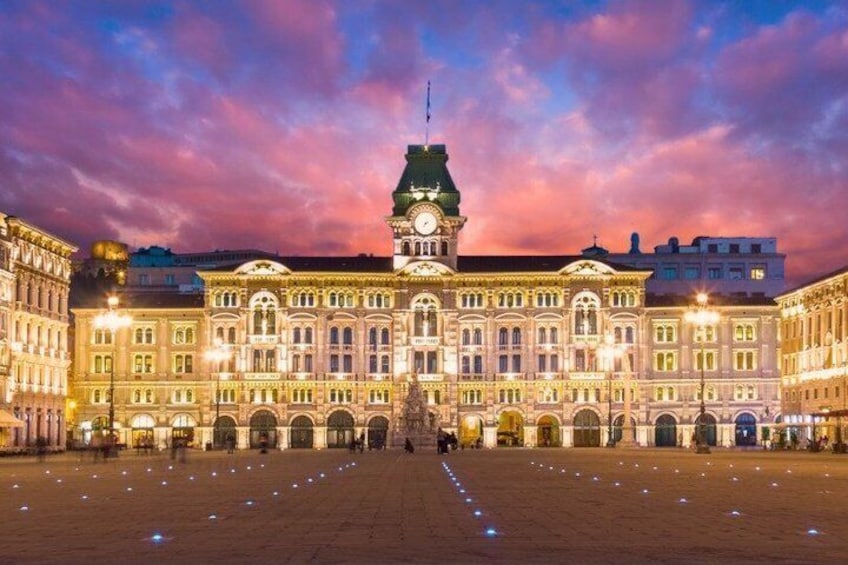 A grand building with a clock tower, illuminated by lights, and a large open plaza. 5