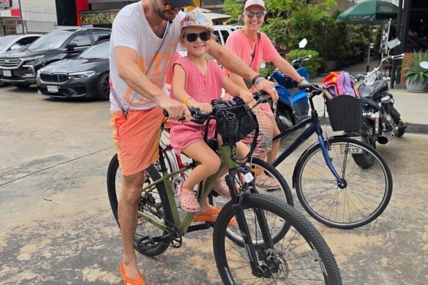 A man and a child riding a bicycle on a street with parked cars and motorcycles in the background. 2