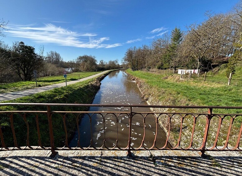 A narrow waterway flanked by grassy banks and trees, with a metal railing in the foreground. 1