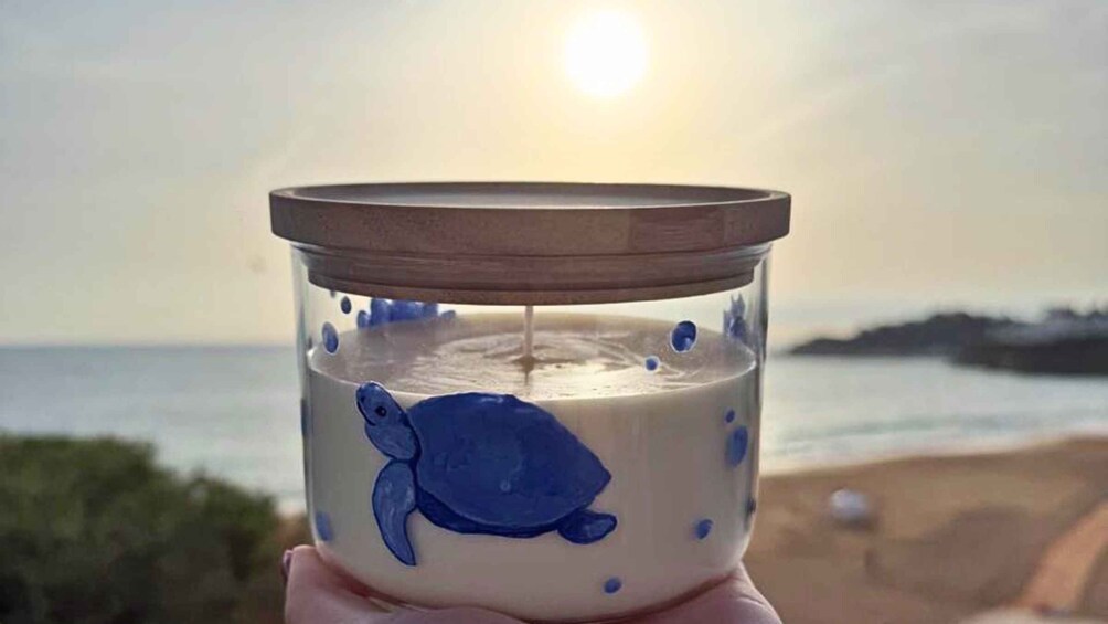 A candle with a turtle design in a glass holder against a beach backdrop with the sun. 2